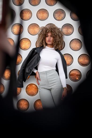 A woman with curly, voluminous blonde-brown hair stands in front of a white wall decorated with symmetrically arranged, circular, orange-lit lamps. She is wearing a fitted white long-sleeve top, high-waisted light gray pants, and a black leather jacket draped loosely over her shoulders. The perspective of the image is framed by blurred black shapes in the foreground, drawing attention to the woman. Her posture is confident, with one hand on her hip and a direct gaze at the camera.
