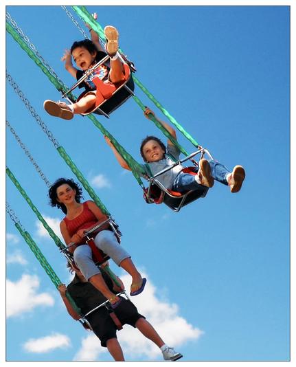 This image shows three people enjoying a ride on a chair swing at an amusement park. They are suspended in the air, each strapped into individual swing seats attached to chains. The sky is clear blue in the background, suggesting a sunny day, perfect for such outdoor activities. The two children and one adult in the image all appear to be having a great time, with their expressions ranging from excited to joyful. The motion of the swing is captured in the angle of the chains and the position of their legs, conveying a sense of movement and enjoyment.