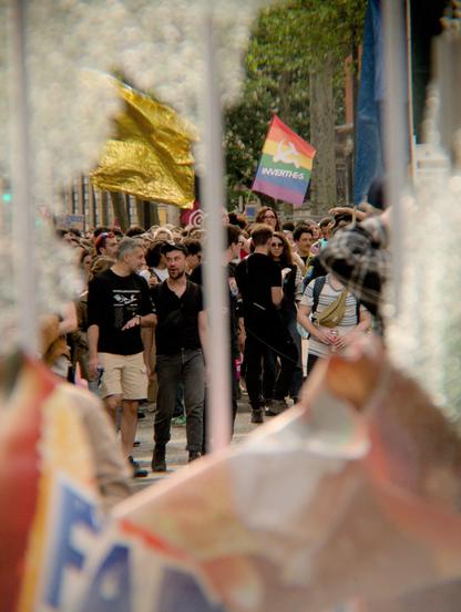 L'image montre une foule de personnes participant à une manifestation. Elles tiennent des drapeaux, dont un drapeau arc-en-ciel avec le texte "inverti·es". le coutour de l'image est floue, la photo est prise à travers les reste d'un panneau publicitaire de verre cassé dont on voit l'affiche publicitaire froissée et tombée sur le bas de l'image.