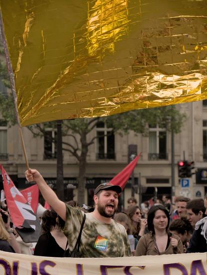 L'image montre le militant Tarik Safraoui tenant un grand drapeau fait d'une couverture de survie dorée lors d'une manifestation. La personne porte une chemise tie-dye et une casquette, et semble crier ou chanter. Il y a d'autres personnes à l'arrière-plan, certaines tenant des drapeaux, et elles sont rassemblées dans une rue avec des bâtiments typiquement parisiens visibles derrière elles.