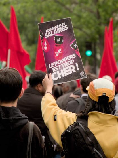L'image montre une personne tenant une pancarte dans une foule de manifestation. La pancarte dit : « AUGMENTEZ LES SALAIRES, LE POPPERS EST TROP CHER ! ». Dans la foule, on voit plusieurs drapeaux rouges en arrière plan.
