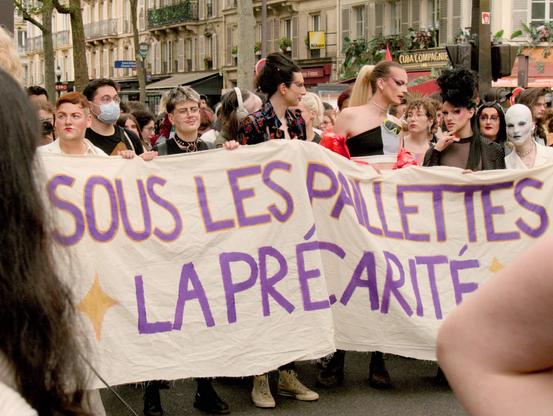 L'image montre un groupe de personnes tenant une banderole lors d'un rassemblement public ou d'une marche. La banderole indique « Sous les paillettes, la précarité ». Les personnes sont vêtues de différentes manières, certaines étant maquillées et costumées en Drag Queen. La scène se déroule dans une rue typiquement parisienne, avec des bâtiments en arrière-plan.