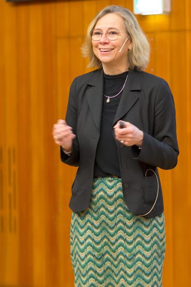 Das Bild zeigt Britta Redlich beim DESY-Nutzertreffen 2025, bei dem sie einen Vortrag hält. Britta ist Chemikerin und Professorin für Experimentalphysik mit umfangreicher Forschungserfahrung. Nach ihrem Studium und ihrer Promotion in Deutschland arbeitete sie in den Niederlanden, wo sie zuletzt den Freie-Elektronen-Laser FELIX und das Hochfeldmagnetlabor HFML an der Radboud-Universität in Nijmegen leitete. Sie hatte mehrere renommierte Positionen inne, unter anderem als Vorstandsmitglied des Niederländischen Physikrats und als Senatorin bei Helmholtz.

Auf dem Foto ist sie mitten in ihrem Vortrag zu sehen, gekleidet in einem eleganten schwarzen Blazer über einem türkis-beigen gemusterten Rock, ergänzt durch einen schwarzen Rollkragenpullover. Sie wirkt engagiert und begeistert, während sie spricht, und ist mit einem Headset-Mikrofon ausgestattet. Das Ambiente umfasst einen warm beleuchteten Hintergrund mit Holzpaneelen, was die akademische und professionelle Atmosphäre unterstreicht.

[ChatGPT 4o]
