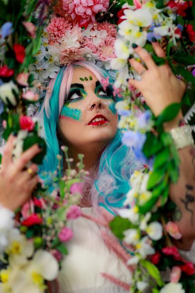 “Portrait of a person with vibrant blue and pink hair, adorned with a floral crown made of pink, white, and colorful flowers. Their makeup is bold, featuring glittery green and blue accents around the eyes and vibrant red lipstick. Surrounded by lush flowers and greenery, they gaze upwards, creating a whimsical and fantasy-inspired atmosphere.”