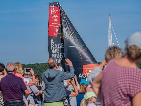 In der Bildmitte sind die Segel der Yacht #TeamMalizia vom OceanRace zu sehen. Auf dem Groß steht "A Race We Must Win" Im Vordergrund sind Zuschauer auf dem Falckensteiner Strand zu sehen, die sich alle dem Boot zu gewendet haben und ihre Telefone hoch halten.