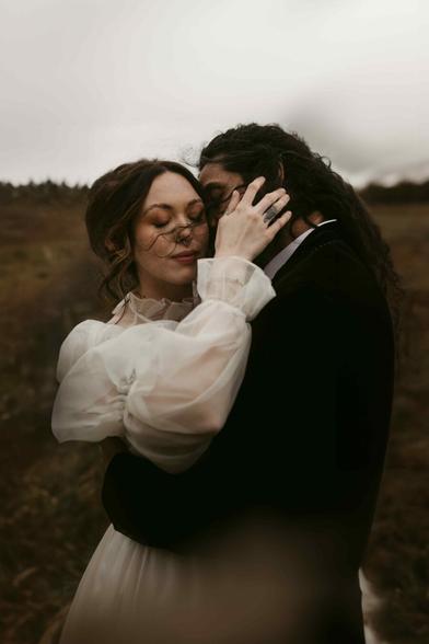 This image shows a couple embracing tenderly. The woman is wearing a long-sleeved white dress made of romantic, sheer fabric that appears soft and flowing. Her face is turned toward the viewer; she is smiling gently with her eyes closed, while a strand of hair is swept across her face by the wind. The man, with long dark hair, is holding her lovingly and seems to be touching her forehead or head. The scene takes place in a natural, slightly barren landscape – the background is blurred, dominated by earthy browns and greens, along with an overcast sky. The atmosphere feels intimate, calm, and emotional. This photograph was captured by European elopement wedding photographer Sturmsucht, known for embracing natural elements and creating moody, timeless imagery.