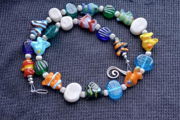 Set against a dark blue background, a handmade lampwork glass bead necklace, featuring fish shaped beads sculpted in the torch flame, and textured beads in a wide range of colors.