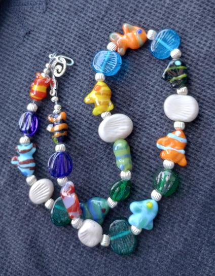 Set against a dark blue background, a handmade lampwork glass bead necklace, featuring fish shaped beads sculpted in the torch flame, and textured beads in a wide range of colors.