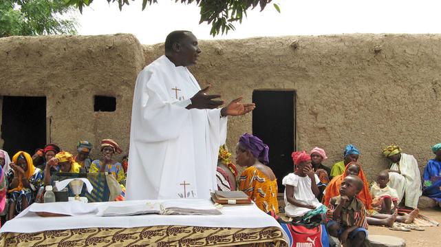 Das Bild zeigt einen Priester in weißen Gewändern, der vor einer Gruppe von Frauen und Kindern in einem einfachen Lehmhaus eine Messe oder eine ähnliche religiöse Zeremonie abhält. Die Frauen tragen bunte Kopfbedeckungen, und die Atmosphäre wirkt friedlich und andächtig. Der Hintergrund deutet auf eine ländliche, möglicherweise afrikanische Umgebung hin.