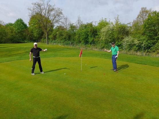 Auf einem Grün auf einem Golfplatz stehen zwei Männliche Golfer. Um die Fahne herum liegen zwei weiße und ein roter Golfball