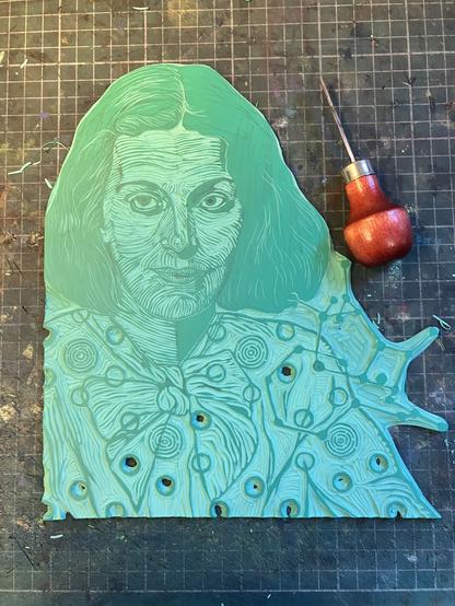 My Lino block (green vinyl) in progress is mostly carved and shows a young Dorothy Crowfoot Hodgkin from chest up in reverse in a blouse tied with a bow. Her ball-and-stick molecular model for penicillin appears over her shoulder and her blouse has a specific repeating pattern of circles and nested broken circles like a labyrinth.