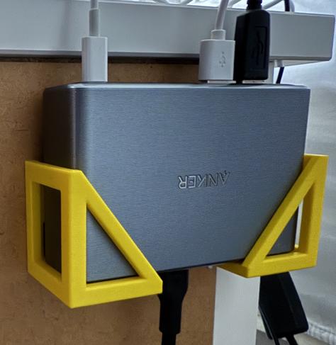 A close-up image of a 3D-printed holder or "basket" designed to secure a rectangular Anker charger. The holder is yellow, with a geometric triangular frame on each side, mounted under a desk or surface. The charger is neatly fitted into the holder, with multiple cables connected to it from the top and bottom.