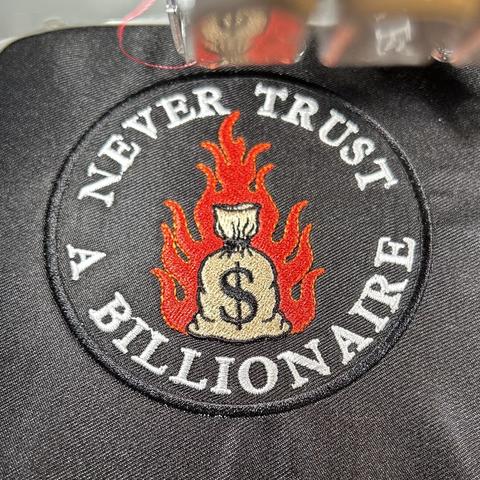 Embroidered black patch with the text “Never Trust A Billionaire” with a flaming bag of money in the center of the patch.