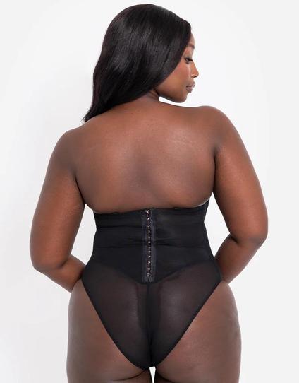 Czarnoskóra modelka w czarnym gorsetowym body bez ramiączek (strapless), zapinanym z tyłu na kilkanaście haftek, stoi plecami do kamery z głową skręconą w bok. Zapięcie zajmuje rejon talii, poniżej jest część majtkowa z półprzejrzystego elastycznego tiulu, wysoko wycięta na biodrach.