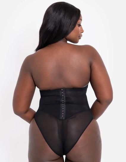 A Black model in a black strapless corset-style bodysuit, fastened at the back with dozens of hook-and-eye closures, stands with her back to the camera and her head turned to the side. The closure covers the waist area, while below is a high-cut panty section made of semi-sheer stretchy tulle.