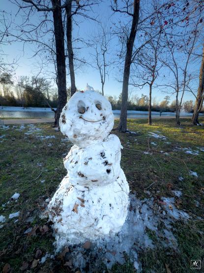Color photo of a snow person with stones for eyes and a curved stick for a mouth. In the background are trees and a small lake.