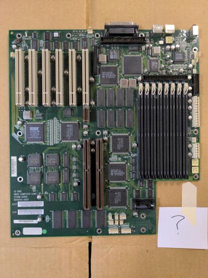 A green computer motherboard lying on cardboard. It has six PCI slots, two ASPD slots, eight RAM sockets, and only a few builtin external ports along the top edge.