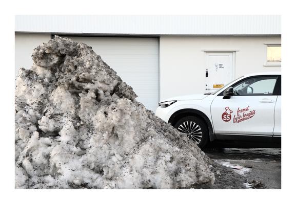 A dirty snow pile sits in front of a white commercial building with a branded car parked beside it. The car displays a red logo and text that translates to "SS, known for taste."