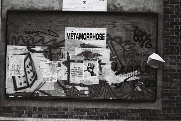 Shoot of ripped off posters and flyers on a brick wall. We can read "metamorphosis" in french and " EU should be defending human rights everywhere" in English. On the left there is a rough sketch of a dice made with spray paint. On the right we can read "colèr-" (anger in French). he rest of the flyers are difficult to decipher and give a chaotic feeling.