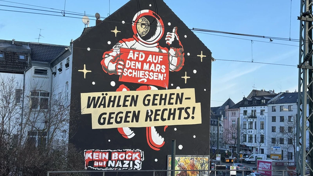 Photo of a panel pained on the side of a house with an astronaut holding a plaque "AFD auf den Mars schiessen" (something like "send AFD to Mars"), with the text "wählen gehen - gegen Rechts" ("go vote - against the right") superimposed.
To the lower left of the panel the text "Kein bock auf Nazis" ("No patience for/interest in Nazis") can be read, which is the name of an anti-racist activist group.