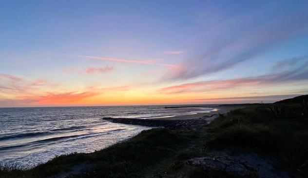 Das Bild zeigt einen Sonnenuntergang über dem Meer. Der Himmel ist in warmen Farbtönen gefärbt, von rosa und orange bis hin zu einem leuchtenden gelb. Die Sonne selbst ist nicht sichtbar, aber ihre Reflexion auf den Wolken und dem Wasser ist deutlich zu erkennen.
Im Vordergrund ist ein grasbewachsener Hügel zu sehen, der sanft zum Meer hin abfällt. Ein felsiger Wellenbrecher erstreckt sich in die Ferne und teilt das Meer in zwei Teile. Die Wellen brechen sanft an der Küste und erzeugen einen leichten Schaum.