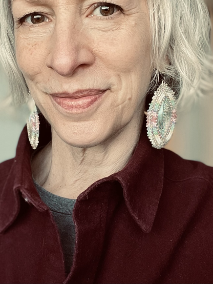 A wrinkled old lady wears beautiful flat beadwork earrings in winter shades of white, silver, blue, and pink.