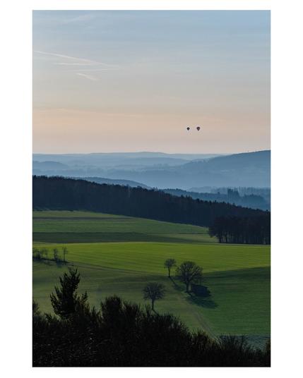 Foto im Hochformat. Blick über die schwäbische Alb von einem hohen Punkt aus. Im Vordergrund sind Wiesen mit vereinzelten Bauern, dann kommen Wälder. Dahinter Berge und Täler in denen der Nebel hängt. Am Himmel sind zwei Heißluftballons relativ nah beieinander. Der Himmel ist blau mit wenigen Wolken.