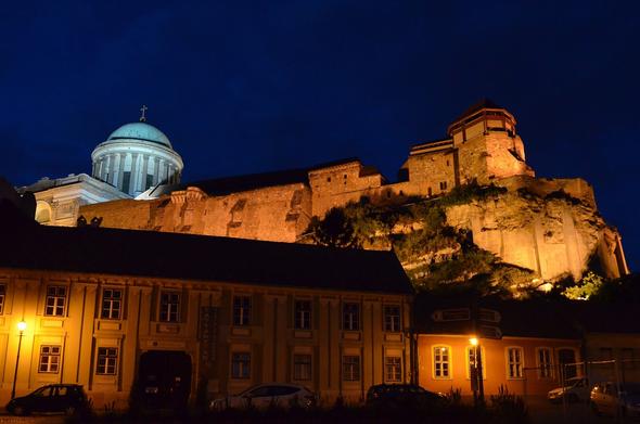 Night view over Esztergom castle with the cathedral in the background, illuminated in yellow with a dark blue evening sky in the background.