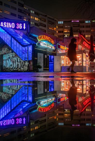 A nighttime cityscape with neon-lit signs. At the center, there is a casino entrance with the text "CASINO 36" in red and yellow letters and a blue glowing arrow. To the right, another illuminated sign reads "BURGER HOUSE" in red. On the left, a staircase with blue-lit glass walls and a wall covered in graffiti can be seen. In the foreground, a puddle reflects the neon signs and surrounding buildings, creating a symmetrical effect. On the right side, the silhouette of a person walking by is visible. In the background, multi-story residential buildings with lit windows are present.