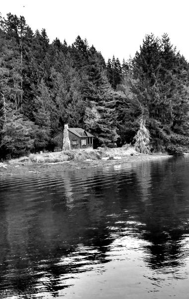 A black-and-white photograph showcasing a rustic cabin situated by a calm river. The cabin features a stone chimney and is nestled against a backdrop of dense evergreen trees. The water reflects the surrounding landscape, adding to the serene atmosphere of this natural setting.