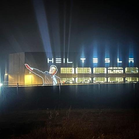 Una proyección nocturna en la gigafábrica de Tesla en Berlín, Alemania, reproduce la imagen reciente de Elon Musk realizando el saludo al estilo nazi, acompañada por el texto iluminado "HEIL TESLA" en la fuente característica de Tesla.