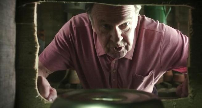 Tom Wilkinson looks through a window like opening. He appears to be talking to someone on the other side.