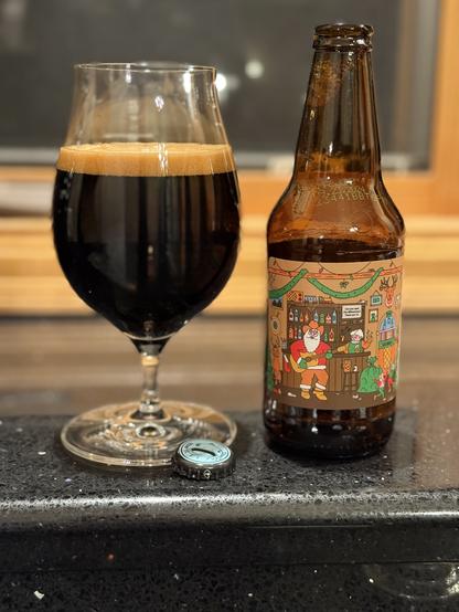 A tall snifter glass of dark beer next to a brown bottle with a colorful label showing Santa in a red suit holding a guitar in a bar festooned with Christmas decor