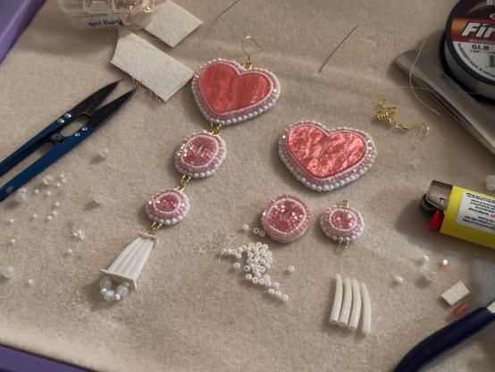 A bead mat with a flat beadwork and dentalium earring project in progress

The earrings feature pink resin heart centers lined with white and pink beads, two beaded circle pieces, and a bottom piece with four dentalium shells and white beads. These components are connected by gold thread guards and jump rings.

You can see blue thread snips, white beads, dentalium shells, faux leather scraps, a yellow lighter, a spool of thread, a bead scooper, a needle, and some gold findings on the bead mat.