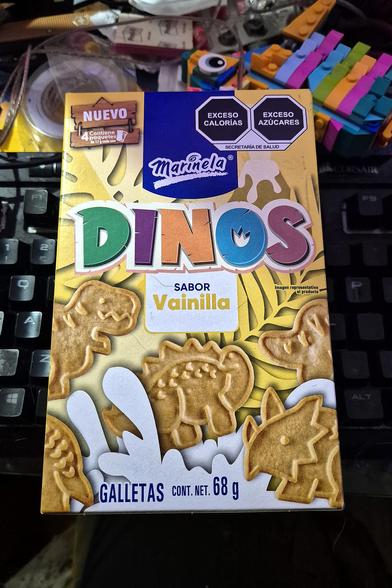 The image shows a box of Marinela Dinos vanilla-flavored cookies. The box is yellow and blue and features illustrations of dinosaurs. The box is sitting on a desk next to a keyboard.