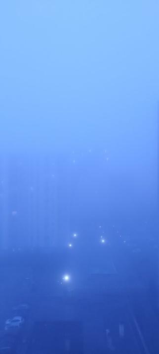 Vue depuis un immeuble en hauteur vers d'autres immeubles. L'image est toute bleue et on ne voit pas grand chose à cause du brouillard, les immeubles au fond sont à peine visibles, quelques lumières font des tâches lumineuses, on distingue des voitures garées