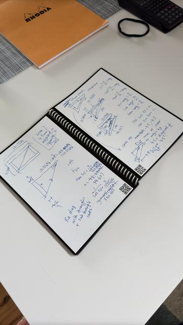 Scribbled out plans and calculations for a triangular -prism shaped enclosure I'm building. They lay on a white desk, in a spiral notebook with black-rimmed white pages. Scribble is in blue ink.