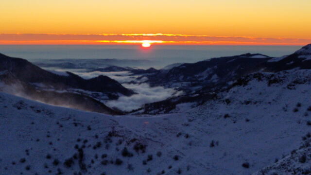 Format paysage. Coucher de soleil depuis une cime enneigée, en regardant vers une vallée partiellement remplie d'une mer de nuages. Le quart haut orangé, les trois quarts bas bleu sombre violacé. La vue donne à travers l'ouverture d'un col enneigé et battu par une bise crépusculaire. De la neige soulevée par le vent se distingue à gauche. Derrière, de part et d'autre, les deux lignes de crêtes des montagnes boisées et sombres plongent vers le fond de vallée et la plaine lointaine. Le brouillard les comble, formant une nappe dont les ondulations sont subtilement relevées par les derniers rayons rasants.