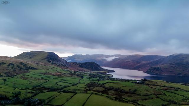 Aerial shot over farmland with Ennerdale Waters lake in distance surrounded by mountains. Heavy clouds in sky