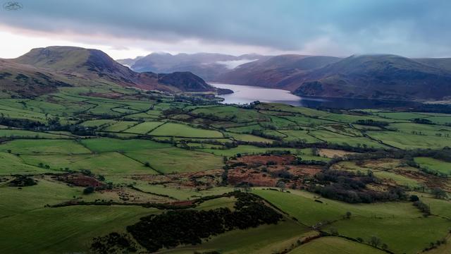 Aerial shot over farmland with Ennerdale Waters lake in distance surrounded by mountains. Heavy clouds in sky