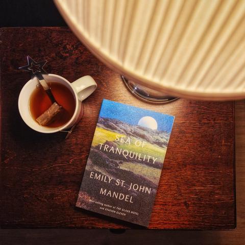Emily St. John Mandelin kirja Sea of Tranquility puisella yöpöydällä pöytälampun ja teekupin vieressä.