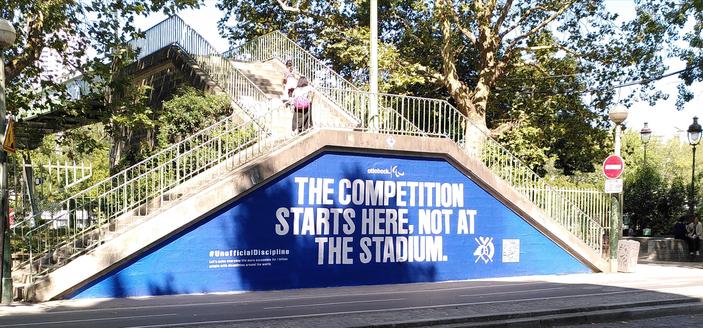 Pignon d'un double escalier urbain permettant d'accéder à une passerelle encadrée par des arbres feuillus. Le pignon est recouvert d'une publicité (pour l'entreprise de fabrication d'orthèses et prothèses) : inscription en blanc "The competition starts here. Not at the stadium", sur fond bleu.