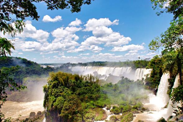 Under a blue sky scattered by white clouds, amidst a lush tropical forest, several waterfalls create a majestic show. Nature at its best in a sequence of greens, whites and blues.