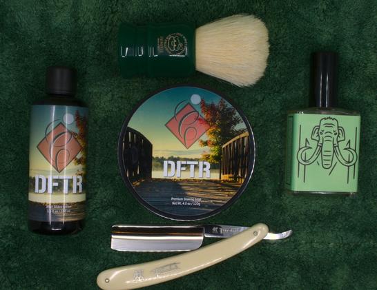(ChatGPT description) 

This image displays a carefully arranged wet shaving setup on a plush green towel, featuring a selection of premium grooming products. At the center is a tub of DFTR Premium Shaving Soap, with a scenic label depicting a sunset over a bridge, symbolizing a calming and nature-inspired shaving experience. To the left, a matching bottle of DFTR After Shave Lotion complements the soap, creating a cohesive set designed for post-shave care.

On the right, a bottle of House of Mammoth Shire Eau de Parfum stands out with its distinctive green packaging and mammoth logo. House of Mammoth is known for its unique and creative scents, and the "Shire" fragrance likely evokes earthy and warm notes.

Above the soap is a Zenith 2022 Brush of the Year, featuring a deep green handle with an engraved logo and natural boar bristles, a popular choice among wet shaving enthusiasts for its excellent lathering capabilities and traditional craftsmanship.

Below the soap is a Henckels Friodur 17 straight razor, recognized for its ivory-colored handle and a stainless steel blade that offers exceptional edge retention and precision. This razor is a testament to the heritage and durability of German craftsmanship.

The combination of these elements creates a visually appealing and functional shaving kit, ideal for those who appreciate both modern artistry and classic grooming traditions. The green background enhances the luxurious and natural aesthetic.