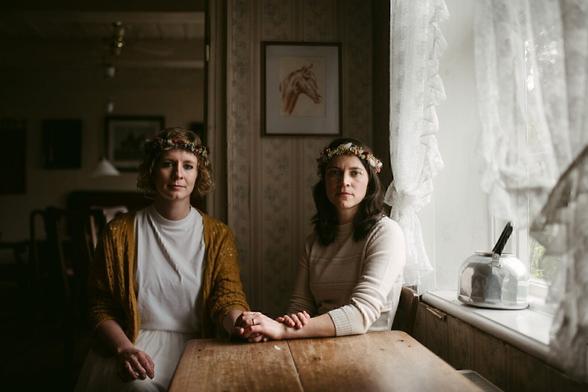 This image captured by Hamburg elopement and wedding photographer Sturmsucht shows an intimate and warm moment between two women seated at a wooden table. Both wear floral crowns, adding a delicate and natural touch to their look. One wears a mustard-yellow cardigan over a white dress, while the other is dressed in a soft cream sweater. They hold hands gently on the table, symbolizing connection and support. The setting is indoors, with soft natural light streaming through lace curtains, creating a nostalgic and calm atmosphere. The background features simple vintage decor, including a framed drawing of a horse and muted wallpaper, complementing the serene and timeless feel of the photograph.
