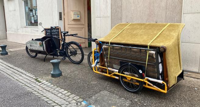 Le vélo-cargo électrique de Mines de Rayons, avec sa remorque attachée à l'arrière, sont stationnés devant le local de l'association. Un gros canapé est solidement sanglé sur la remorque. Ça donne l'impression que la plus grosse difficulté n'est pas le transport à vélo, mais le passage de la porte cochère !