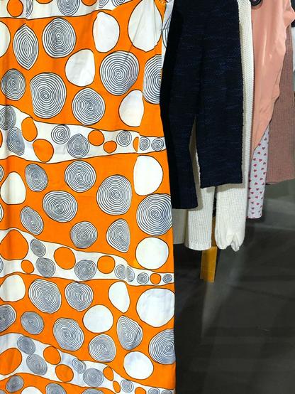 On the left half of the frame is a large section of a bright late 60s/ early 70s long ornage and white dress featuring many circles (some with spirals) next to each other. On the right side of the frame are some boring dark clothes on a rack.