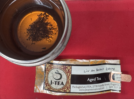 A steel thermos of freshly brewed tea. Next to it a packet of the tea “Liu A Basket 2014” from the sellers “jteainternational.com” the tea is a luminous golden brown and some bits of leaves have drifted to the bottom.