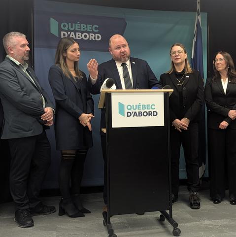 Claude Villeneuve, en complet cravate, se tient derrière un pupitre où l'on peut lire Québec D'abord. Des micros y sont déposés. À sa gauche deux personnes, à sa droite, deux autres personnes.