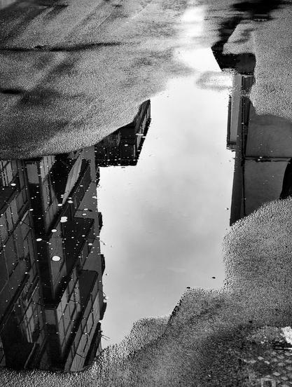 Foto in bianco e nero di una pozzanghera di acqua piovana che riflette un palazzo e una porzione di cielo grigio